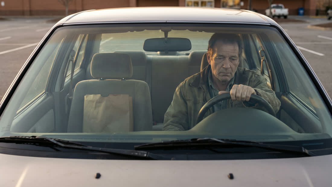 A man in his late fifties sits alone in a parked car in warm afternoon light, hands on the wheel, taking a quiet moment before going back inside.